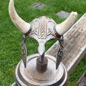 Puede incluir: Un casco vikingo de plata y oro con cuernos sobre un soporte de metal oxidado. El casco tiene un dise&ntilde;o de nudo celta.