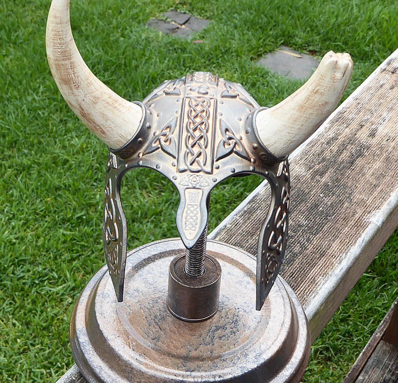 Puede incluir: Un casco vikingo de plata y oro con cuernos sobre un soporte de metal oxidado. El casco tiene un dise&ntilde;o de nudo celta.