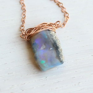 May include: A necklace featuring a rectangular, dark-toned opal pendant with iridescent blue and purple flashes. The stone is secured by a copper-coloured wire wrap at the top, which connects to a matching copper-coloured metal chain link necklace.