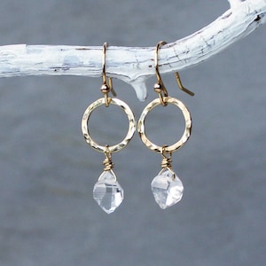 May include: A pair of gold-toned earrings featuring a hammered circular hoop design. Each earring suspends a small, clear, raw-cut crystal gemstone attached with delicate gold wire wrapping, hanging from a simple gold-toned ear wire hook.