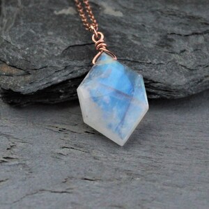 May include: A necklace featuring a faceted, diamond-shaped moonstone pendant with a translucent, iridescent blue glow. The gemstone is secured by a copper wire wrap and hangs from a delicate, matching copper chain, creating a refined look.