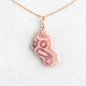 May include: A delicate necklace featuring a thin rose gold chain and a single, irregular-shaped pendant made of pink rhodochrosite stone. The stone displays natural concentric circular banding patterns in shades of light and dark pink.