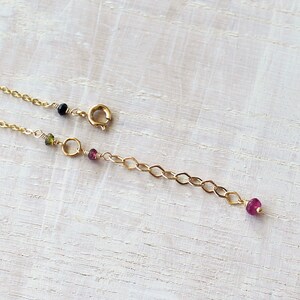 May include: A gold chain necklace featuring a circular spring ring clasp, a short extender chain, and small, multi-colored gemstone beads in shades of black, green, and pink attached at intervals along the delicate metal links.