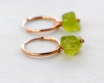Könnte beinhalten: Ein Paar Creolen aus Roségold, wobei jeder Ohrring mit einem einzelnen, rohen und ungeschliffenen limettengrünen Edelsteinanhänger am runden Metallband versehen ist. Die Steine haben eine raue, natürliche Textur und sind mit einer kleinen Metallperle gesichert.