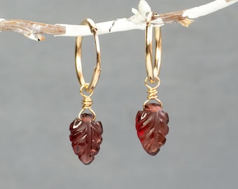 May include: A pair of gold hoop earrings featuring dangling dark red garnet charms carved into the shape of small leaves. Each charm is securely attached to the thin gold hoop with a delicate gold wire wrap, creating a refined and elegant look.