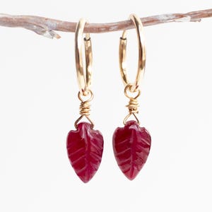 May include: A pair of gold hoop earrings featuring dangling, carved deep red leaf-shaped gemstones. Each gemstone is securely attached to the thin gold hoop by a delicate gold wire wrap, creating a balanced and symmetrical jewelry design.