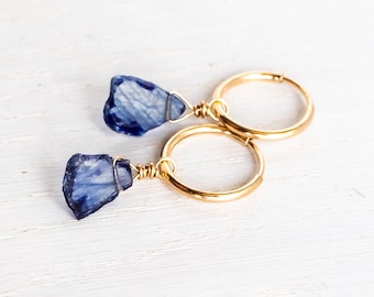 May include: A pair of gold hoop earrings featuring raw, deep blue kyanite gemstone pendants. Each stone is secured to the circular gold hoop with thin gold wire wrapping, showcasing the natural, irregular texture and faceted edges of the crystals.