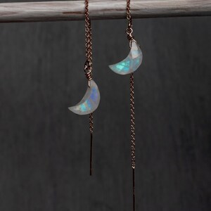 Puede incluir: Un par de pendientes de hilo con cadenas de oro rosa y colgantes de piedra lunar en forma de media luna. Cada pendiente tiene un fino poste de metal en el extremo de la cadena, con el dije de piedra lunar iridiscente suspendido del eslabón.