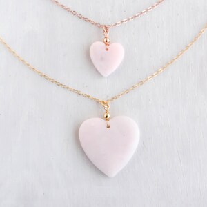 May include: Two layered necklaces featuring smooth, pale pink heart-shaped pendants. The top necklace has a rose gold-tone chain with a smaller heart, while the bottom necklace features a gold-tone chain with a larger heart pendant.