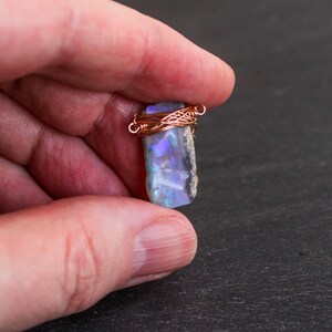 May include: A raw, rectangular opal gemstone featuring iridescent flashes of blue and purple light. The stone is secured at the top with a decorative wrap of thin, metallic copper wire, forming small loops on each side for attachment as a pendant.