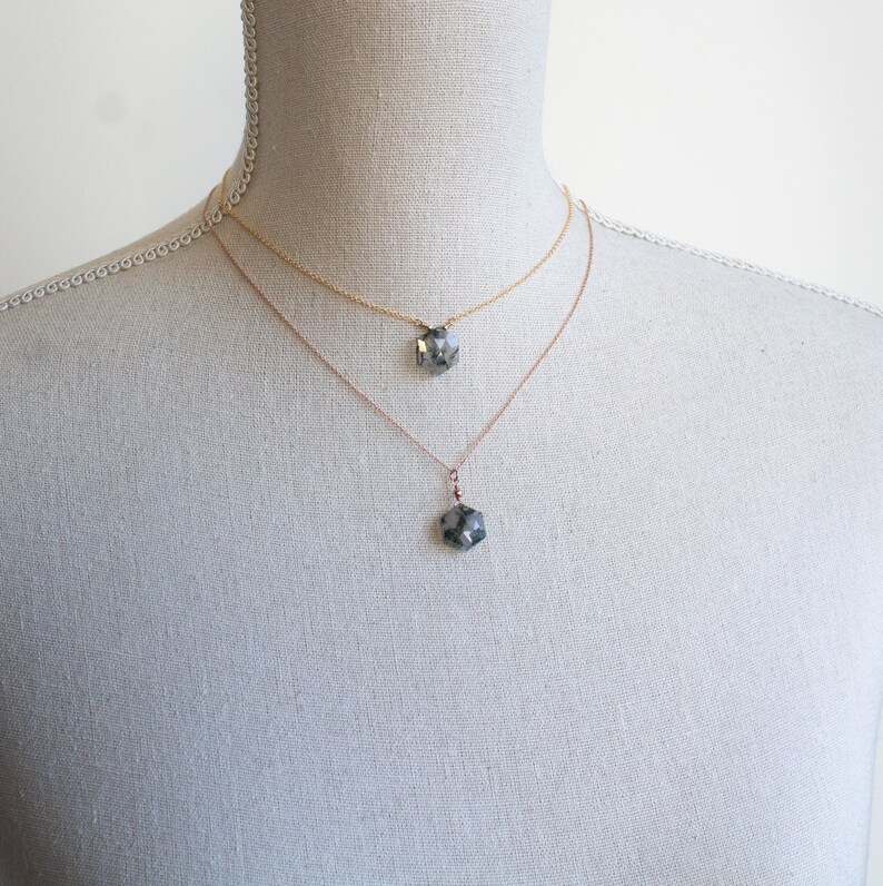 May include: Two layered necklaces featuring delicate gold-toned chains, each holding a single faceted, hexagonal dark gray pendant. The shorter chain sits higher on the neck, while the longer chain features a matching pendant hanging lower.