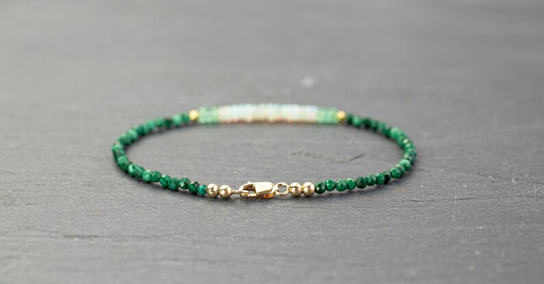 Puede incluir: Una pulsera delicada con peque&ntilde;as cuentas facetadas de color verde oscuro y una fila central de cuentas blanquecinas iridiscentes. El dise&ntilde;o est&aacute; acentuado con diminutas esferas met&aacute;licas doradas y un cierre de mosquet&oacute;n dorado.