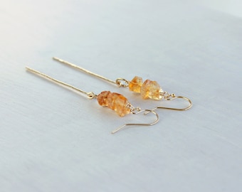 May include: A pair of gold-toned drop earrings featuring long, thin metal bars and small, raw, amber-colored citrine gemstones. The earrings are attached to curved gold-toned ear wires, creating a dangling design with a polished finish.