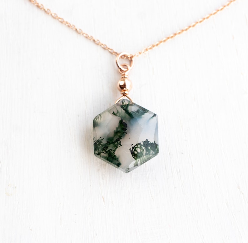May include: A hexagonal moss agate pendant featuring translucent white stone with dark green dendritic inclusions, suspended from a delicate rose gold chain by a small round bead connector. The gemstone has a faceted surface and polished finish.