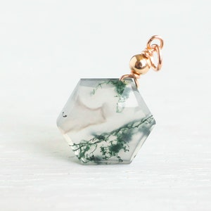 May include: A hexagonal moss agate pendant featuring translucent gray stone with dark green dendritic inclusions. The gemstone is topped with a polished rose gold metal bead and a circular jump ring, creating a delicate and refined jewelry component.