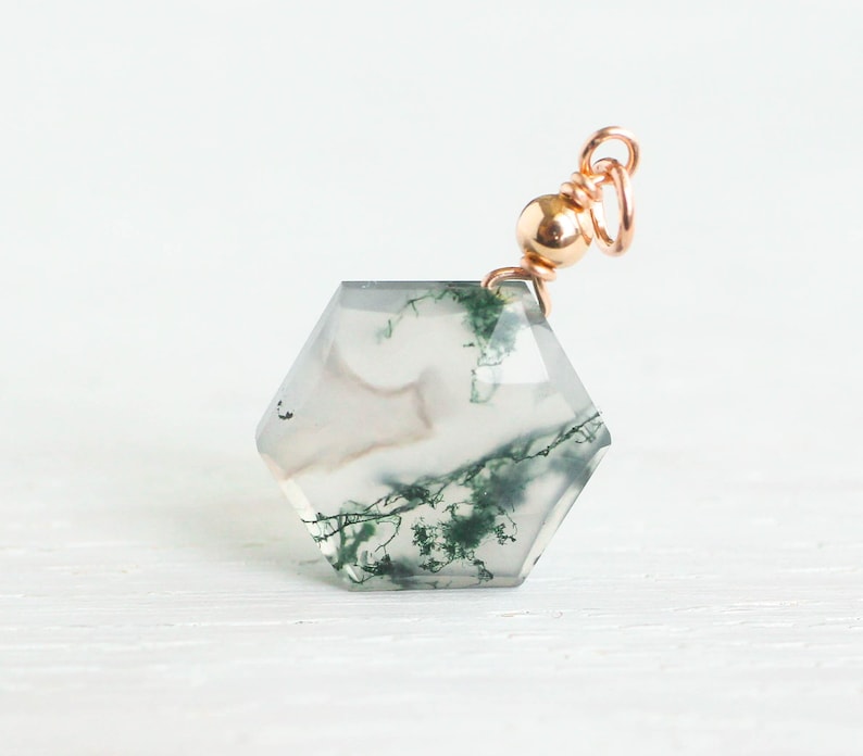 May include: A hexagonal moss agate pendant featuring translucent gray stone with dark green dendritic inclusions. The gemstone is topped with a polished rose gold metal bead and a circular jump ring, creating a delicate and refined jewelry component.