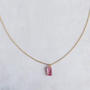 K&ouml;nnte beinhalten: Eine zarte goldene Halskette mit einem einzelnen, kleinen, rechteckigen rosa Edelsteinanh&auml;nger. Der Anh&auml;nger h&auml;ngt mittig an der d&uuml;nnen, polierten, goldfarbenen Metallkette, die einen einfachen, eleganten Bogen vor dem Hintergrund bildet.