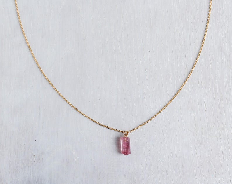 K&ouml;nnte beinhalten: Eine zarte goldene Halskette mit einem einzelnen, kleinen, rechteckigen rosa Edelsteinanh&auml;nger. Der Anh&auml;nger h&auml;ngt mittig an der d&uuml;nnen, polierten, goldfarbenen Metallkette, die einen einfachen, eleganten Bogen vor dem Hintergrund bildet.