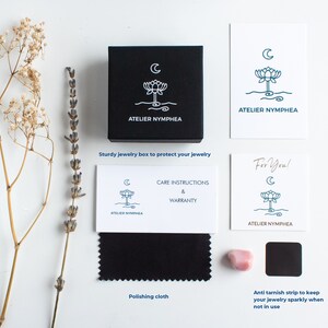 Puede incluir: Una caja de joyer&iacute;a cuadrada negra con un logotipo blanco de loto y luna, acompa&ntilde;ada de tarjetas blancas con el logotipo de Atelier Nymphea, un pa&ntilde;o de pulido negro, una peque&ntilde;a piedra rosa y una tira antideslustre negra, junto a flores secas.