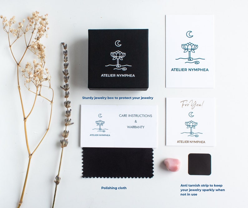 Puede incluir: Una caja de joyer&iacute;a cuadrada negra con un logotipo blanco de loto y luna, acompa&ntilde;ada de tarjetas blancas con el logotipo de Atelier Nymphea, un pa&ntilde;o de pulido negro, una peque&ntilde;a piedra rosa y una tira antideslustre negra, junto a flores secas.