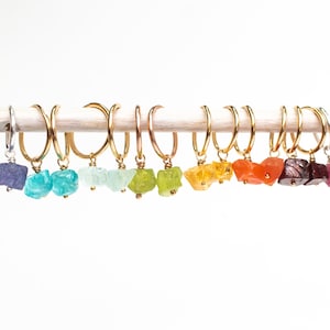 May include: A set of 12 pairs of gold hoop earrings with a variety of colorful gemstone charms. The charms are in shades of purple, blue, green, yellow, orange, red, and pink.