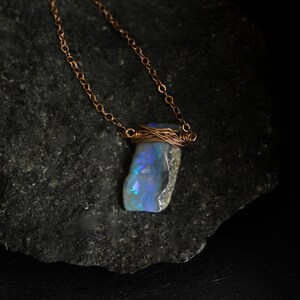 May include: A necklace featuring a raw, iridescent opal pendant secured with copper wire wrapping. The pendant displays flashes of blue and green light against a grey base. It hangs from a delicate copper chain with small, circular links.