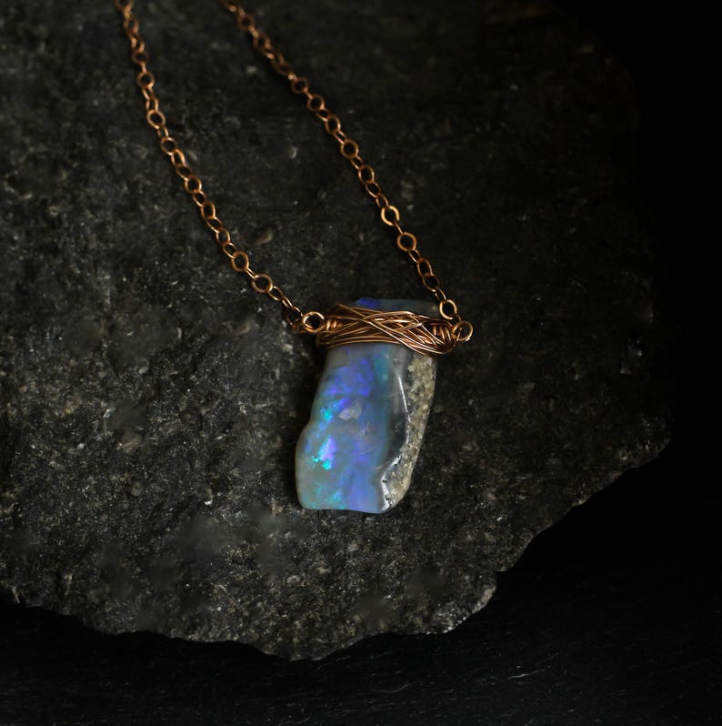 May include: A necklace featuring a raw, iridescent opal pendant secured with copper wire wrapping. The pendant displays flashes of blue and green light against a grey base. It hangs from a delicate copper chain with small, circular links.