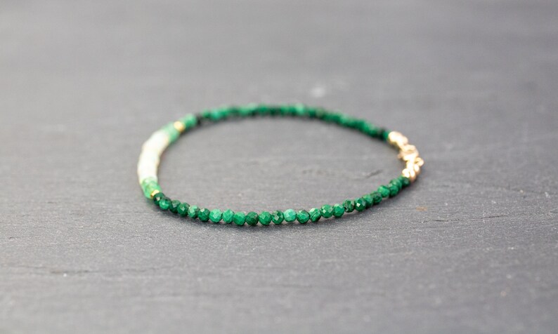 Puede incluir: Una delicada pulsera de cuentas con peque&ntilde;as piedras facetadas de color verde oscuro, una secci&oacute;n corta de cuentas de color crema claro y un peque&ntilde;o cierre de metal dorado. Las cuentas est&aacute;n dispuestas en c&iacute;rculo, mostrando un degradado.