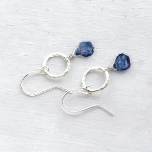 Puede incluir: Un par de pendientes colgantes de plata con un aro circular martillado y una cuenta de piedra preciosa en bruto de color azul intenso suspendida en la parte superior de cada aro. Los pendientes tienen ganchos de plata curvados.