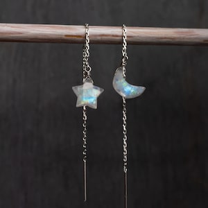 Puede incluir: Un par de pendientes de cadena de plata con un dije de luna y estrella hechos de piedra de luna iridiscente.