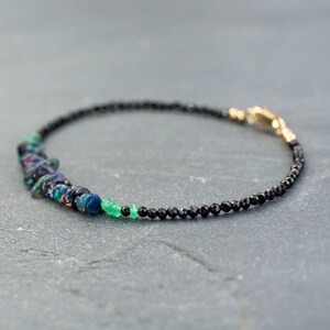 Puede incluir: Una pulsera delicada que presenta una hilera de peque&ntilde;as cuentas negras facetadas, acentuada con un grupo de fragmentos de &oacute;palo iridiscente en azul oscuro y verde, junto a peque&ntilde;as cuentas de piedras preciosas verde brillante y un cierre dorado.