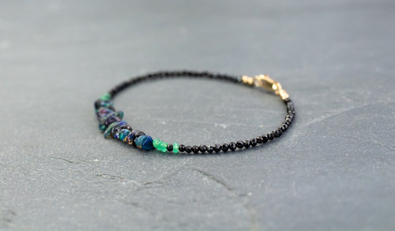 Puede incluir: Una pulsera delicada que presenta una hilera de peque&ntilde;as cuentas negras facetadas, acentuada con un grupo de fragmentos de &oacute;palo iridiscente en azul oscuro y verde, junto a peque&ntilde;as cuentas de piedras preciosas verde brillante y un cierre dorado.