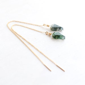 Może przedstawiać: Para złotych kolczyków z zielono-białymi, fasetowanymi zawieszkami z kamieni szlachetnych. Kolczyki są prezentowane na białej powierzchni, podkreślając delikatny łańcuszek i unikalny kształt kamieni.