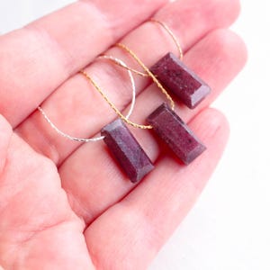 Pode incluir: Close-up de quatro contas retangulares facetadas de pedras preciosas vermelho-escuras, enfiadas em delicadas correntes de ouro e prata. As contas são seguradas na mão de uma pessoa, mostrando sua cor e forma. O fundo é de um tom suave e neutro.