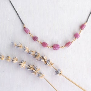 Op de afbeelding: Een delicate ketting met een zilveren ketting en roze edelsteen kralen. De ketting wordt getoond op een wit oppervlak met gedroogde lavendeltakjes.