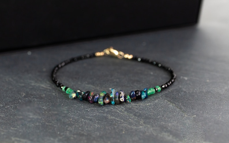 Puede incluir: Una pulsera delicada que presenta una hilera de peque&ntilde;as cuentas negras facetadas, acentuada con una fila central de fragmentos de &oacute;palo iridiscente en bruto y peque&ntilde;as cuentas de piedras preciosas verdes, con cierre de mosquet&oacute;n dorado.