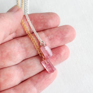 K&ouml;nnte beinhalten: Zwei Halsketten mit kleinen, rechteckigen, durchscheinenden rosa Kristallanh&auml;ngern. Ein Anh&auml;nger h&auml;ngt an einer goldfarbenen Metallkette, der andere an einer silberfarbenen Metallkette. Die Kristalle haben ein facettiertes, poliertes Aussehen.