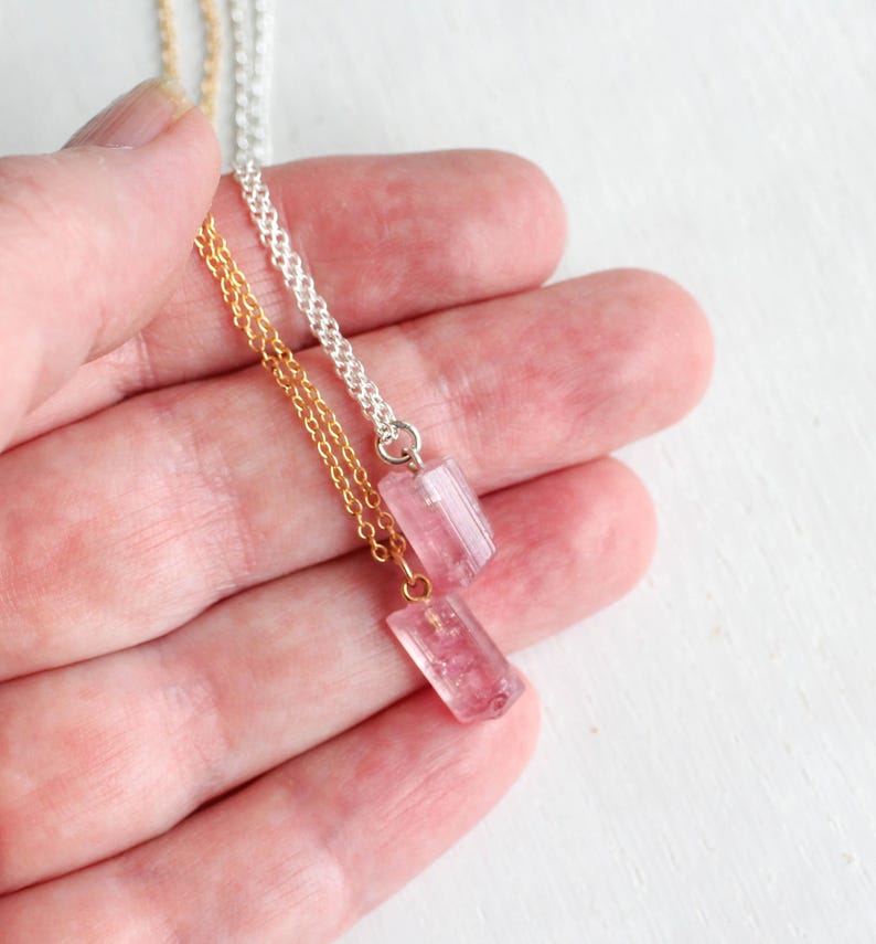 K&ouml;nnte beinhalten: Zwei Halsketten mit kleinen, rechteckigen, durchscheinenden rosa Kristallanh&auml;ngern. Ein Anh&auml;nger h&auml;ngt an einer goldfarbenen Metallkette, der andere an einer silberfarbenen Metallkette. Die Kristalle haben ein facettiertes, poliertes Aussehen.