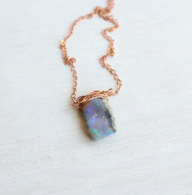 May include: A necklace featuring a rectangular, dark-toned opal pendant with iridescent blue and purple flashes. The stone is secured by a copper-coloured wire wrap at the top, which connects to a matching copper-coloured metal chain link necklace.