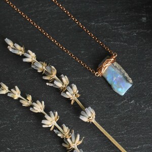 May include: A necklace featuring a rectangular, iridescent blue and white stone pendant wrapped in thin copper wire, suspended from a delicate copper chain. The pendant displays flashes of light and is positioned next to a dried sprig of lavender.