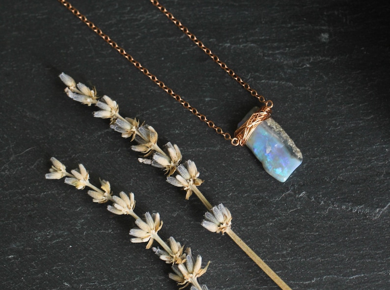 May include: A necklace featuring a rectangular, iridescent blue and white stone pendant wrapped in thin copper wire, suspended from a delicate copper chain. The pendant displays flashes of light and is positioned next to a dried sprig of lavender.