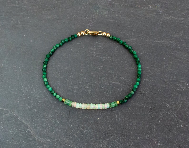 Puede incluir: Una delicada pulsera de cuentas con peque&ntilde;as cuentas facetadas de malaquita verde oscuro y una fila central de cuentas rondelle p&aacute;lidas e iridiscentes similares al &oacute;palo. La pulsera tiene un cierre de mosquet&oacute;n dorado y peque&ntilde;as cuentas espaciadoras doradas.
