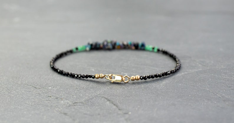 Puede incluir: Una pulsera delicada que presenta una hilera de peque&ntilde;as cuentas negras facetadas, acentuada con un grupo central de fragmentos de piedra azul oscuro y verde. La pieza se completa con un cierre de mosquet&oacute;n dorado y peque&ntilde;as cuentas espaciadoras doradas.