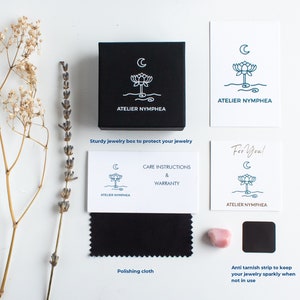 May include: A black square jewelry box with a white lotus and moon logo, accompanied by white cards featuring the Atelier Nymphea logo, a black polishing cloth, a small pink stone, and a black anti-tarnish strip, all arranged with dried flowers.