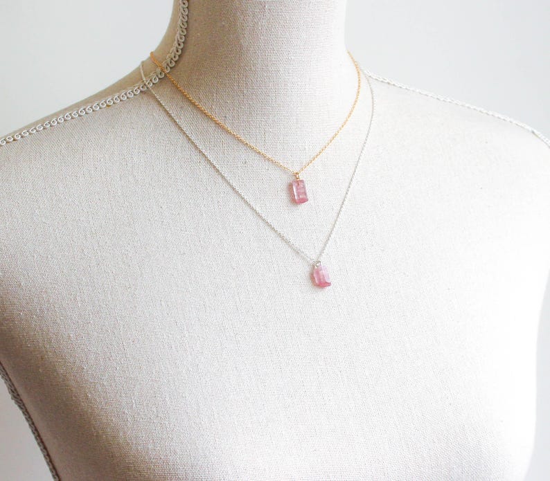K&ouml;nnte beinhalten: Zwei &uuml;bereinander getragene Halsketten an einer Schaufensterpuppe. Die obere Kette besteht aus einer goldfarbenen Kette mit einem kleinen, rechteckigen, durchscheinenden rosa Anh&auml;nger. Die untere Kette hat eine silberne Kette mit einem passenden Anh&auml;nger.