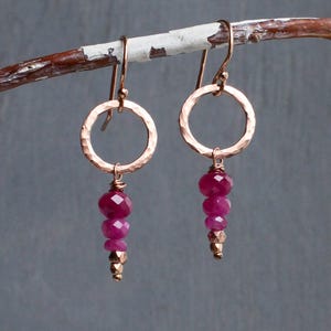May include: A pair of drop earrings featuring a hammered rose gold circular hoop suspended from a hook. Each hoop holds a vertical drop of three faceted deep pink ruby-coloured beads followed by a small metallic bead at the very bottom tip.