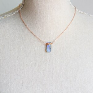 May include: A delicate necklace featuring a thin rose gold chain and a single rectangular pendant made of a translucent, iridescent blue stone. The stone is secured at the top with a decorative rose gold wire wrap that connects to the chain.