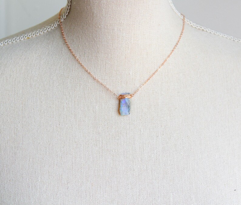 May include: A delicate necklace featuring a thin rose gold chain and a single rectangular pendant made of a translucent, iridescent blue stone. The stone is secured at the top with a decorative rose gold wire wrap that connects to the chain.