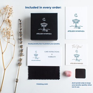 May include: A black jewellery box, a white card with the text ATELIER NYMPHEA, a care instructions card, a For You card, a black polishing cloth, a small pink stone, and a black anti-tarnish strip, all featuring a lotus and moon logo.