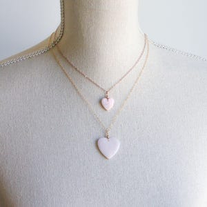 May include: A layered necklace set featuring two delicate rose gold chains, each adorned with a smooth, pale pink heart-shaped pendant. The upper chain holds a smaller heart, while the lower, longer chain features a larger heart pendant.